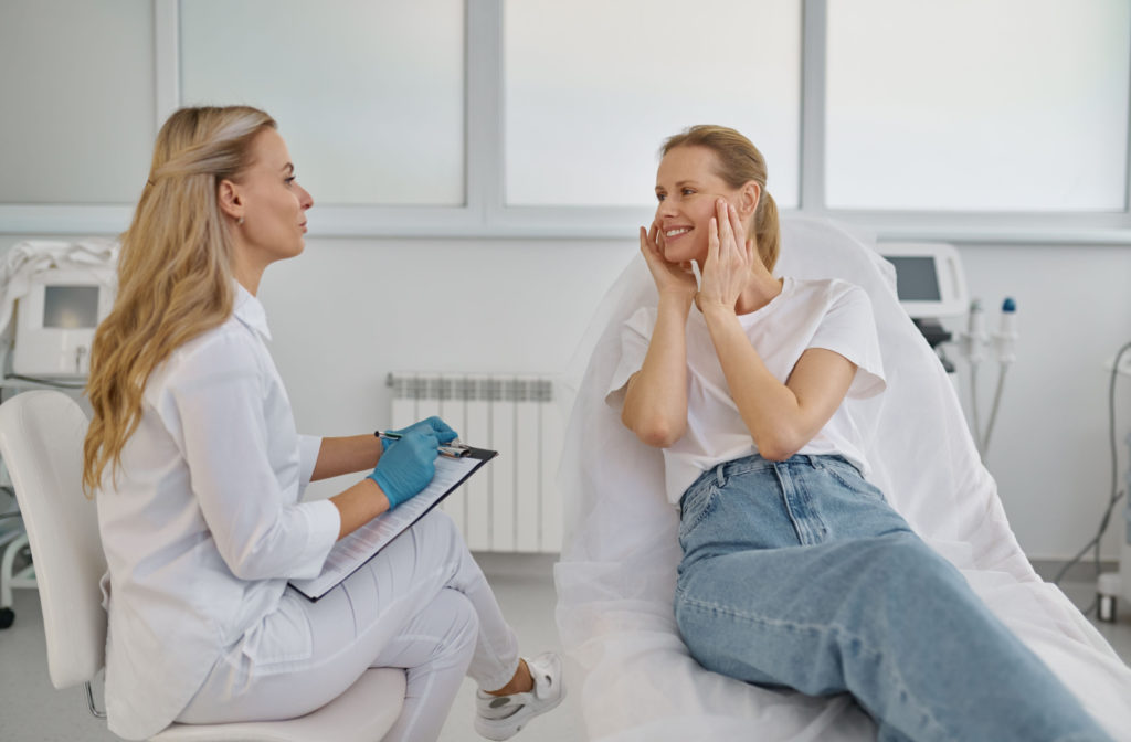 A woman smiling and holding her face with both hands as she sits on a medical bed and looks directly at her doctor, who is holding a clipboard.