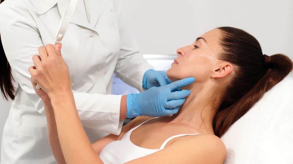 Young woman in an aesthetic clinic looking at her skin in a handheld mirror as a doctor with blue gloves points to a spot on her jawline.