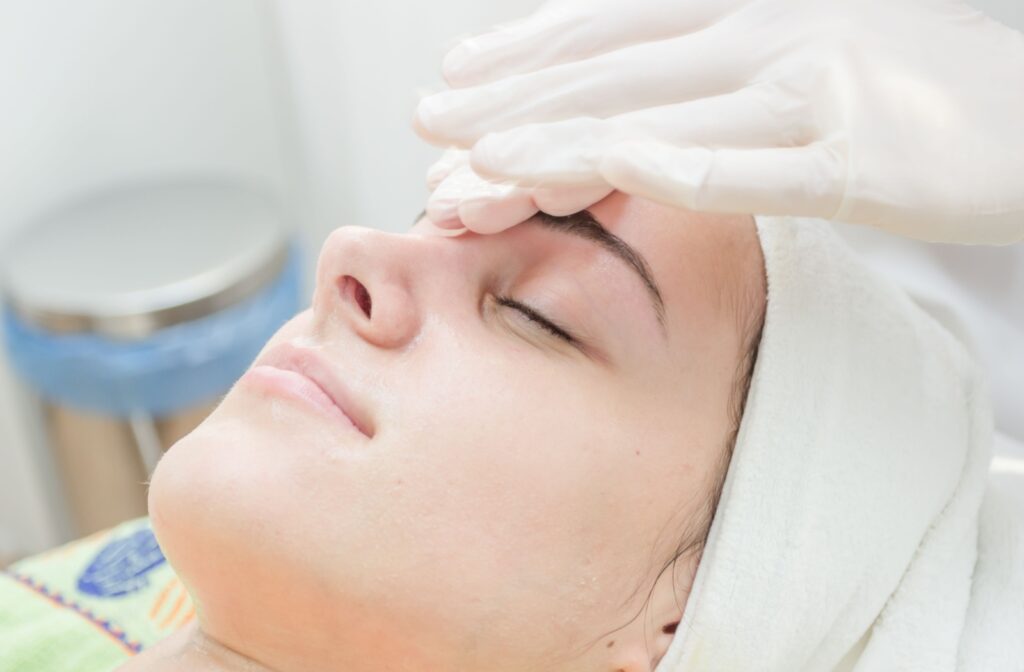 A young woman laying in a spa-like room with her eyes closed as a professional massages her forehead.