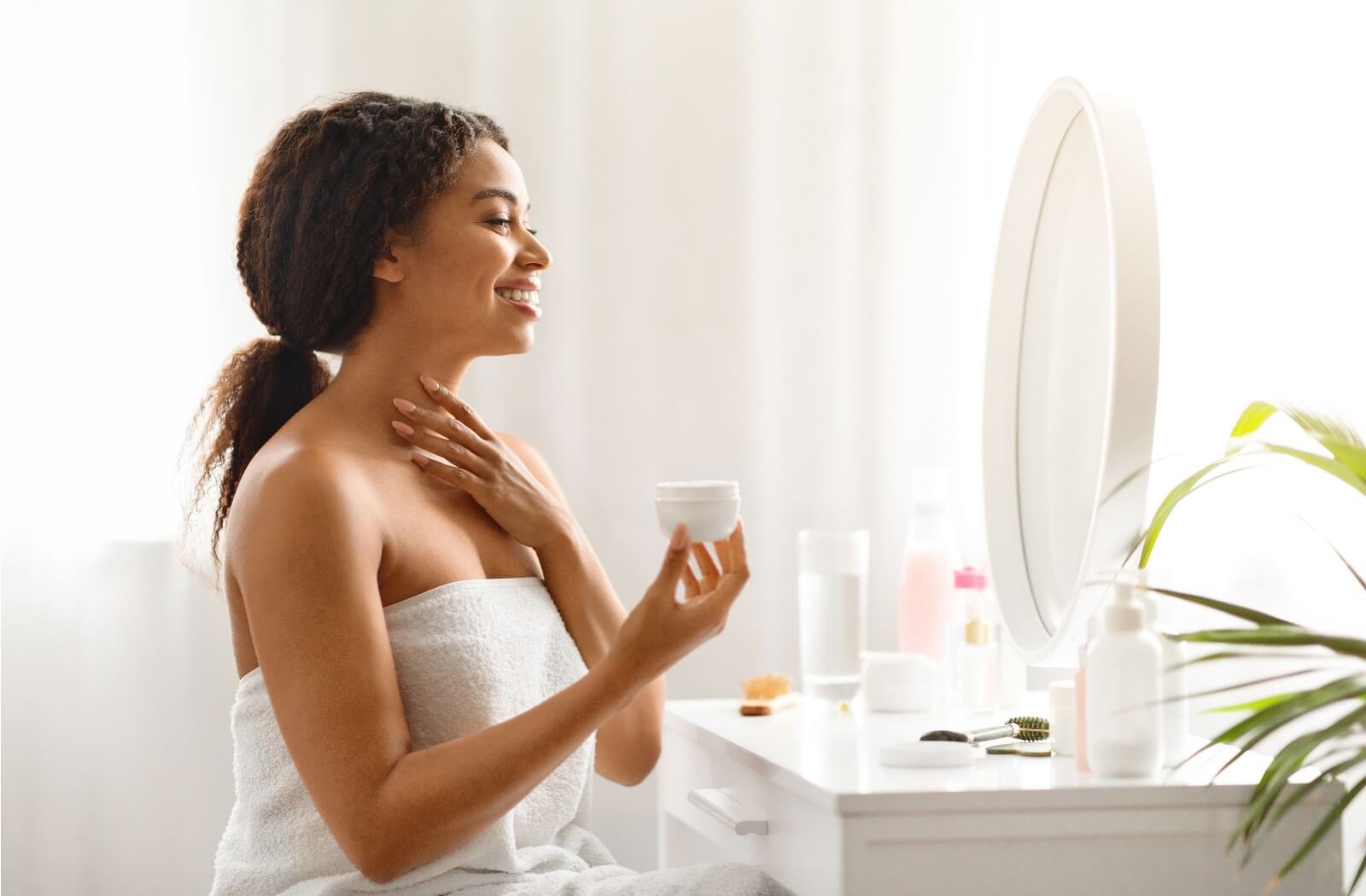Person sitting in front of a vanity in a white towel and applying moisturizer to their neck.